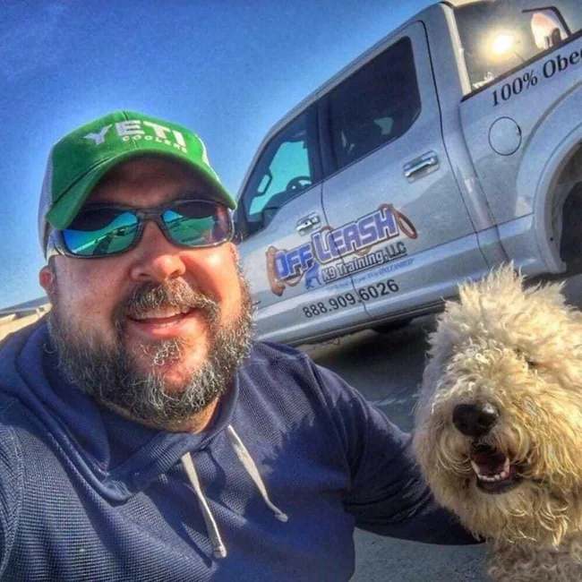 Josh Wilson, owner of Off Leash K9 Training, smiles for a selfie with a shaggy dog in front of the company truck, serving the Charlottesville, VA area.
