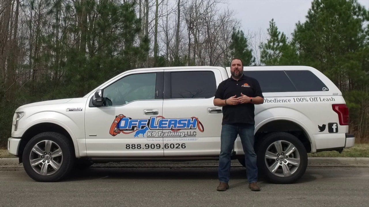 A man stands in front of an Off Leash K9 Training truck, speaking to the camera in a video still promoting the company's 100% off-leash obedience training near Charlottesville, VA.